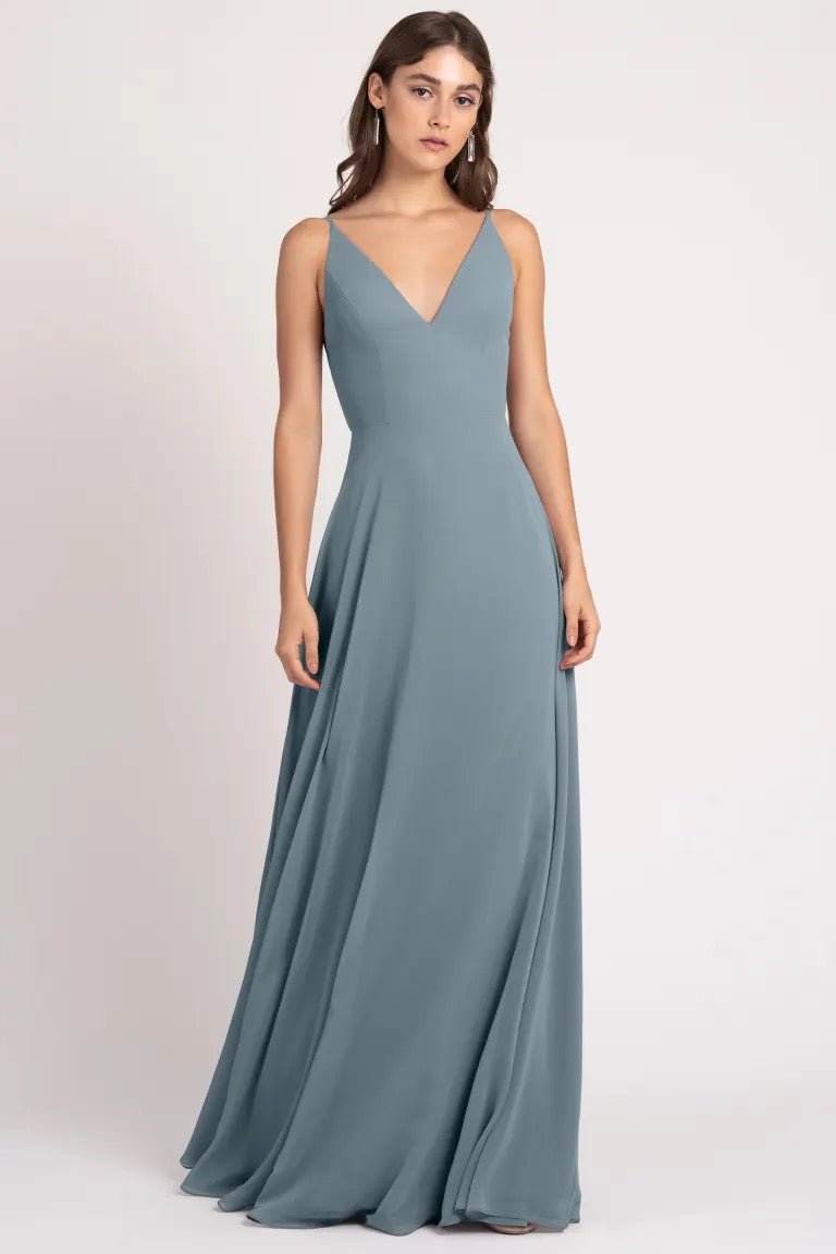 A person wearing a long, slate blue evening gown with sharp tailoring and a v-neckline, Dani - Bridesmaid Dress by Jenny Yoo from Bergamot Bridal.
