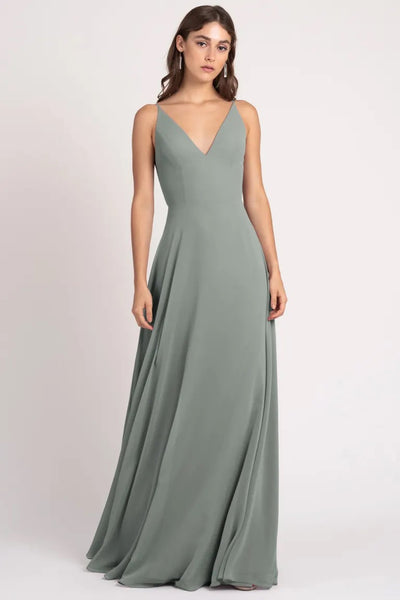 A woman wearing a Dani bridesmaid dress by Jenny Yoo in sage green with a v-line neckline, spaghetti straps, and an a-line silhouette from Bergamot Bridal.