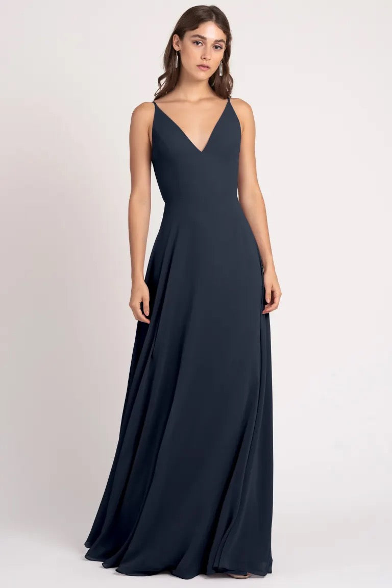 Woman posing in a sleek, navy blue evening gown with an A-line silhouette and a v-neckline, the Dani Bridesmaid Dress by Jenny Yoo from Bergamot Bridal.