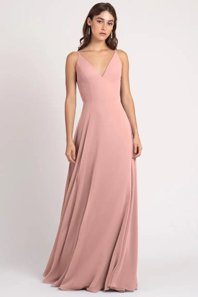 A woman in a pink evening gown with a v-neckline and keyhole back, posing against a neutral background wearing the Dani Bridesmaid Dress by Jenny Yoo from Bergamot Bridal.