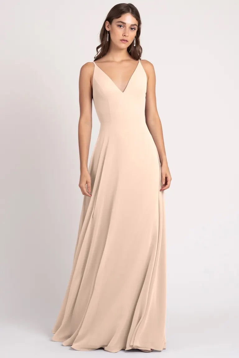 A woman in a sleek, beige Dani bridesmaid dress by Jenny Yoo featuring sharp tailoring and a v-neckline posing against a light background.