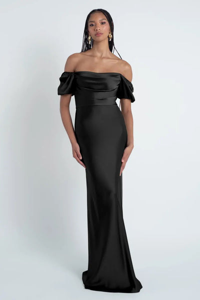 A woman in an elegant black off-the-shoulder Jenny Yoo Bridesmaid Dress with a fitted silhouette and a bias cut skirt in luxe satin fabric from Bergamot Bridal.