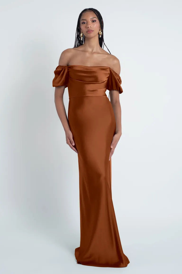 A woman wearing an elegant off-the-shoulder brown Eliana - Jenny Yoo Bridesmaid Dress gown with a bias-cut skirt from Bergamot Bridal.