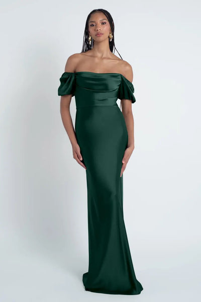 A woman in an elegant green off-shoulder Jenny Yoo Bridesmaid Dress crafted from luxe satin fabric stands against a plain background.