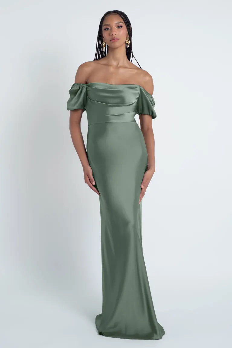 A woman posing in an elegant Jenny Yoo Bridesmaid Dress featuring a luxe satin fabric and an off-the-shoulder green gown from Bergamot Bridal.