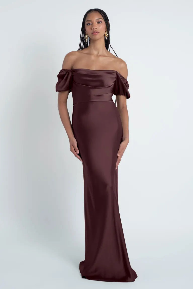 A woman in an elegant off-the-shoulder Jenny Yoo Bridesmaid Dress with a bias cut skirt, standing against a plain background from Bergamot Bridal.
