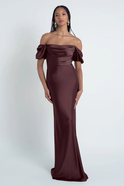 A woman in an elegant off-the-shoulder Jenny Yoo Bridesmaid Dress with a bias cut skirt, standing against a plain background from Bergamot Bridal.