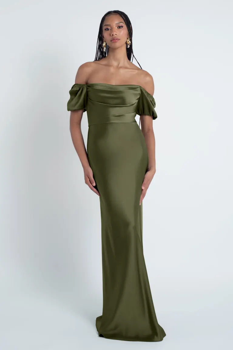 A woman in an elegant olive green off-the-shoulder Jenny Yoo Bridesmaid Dress stands against a neutral background from Bergamot Bridal.