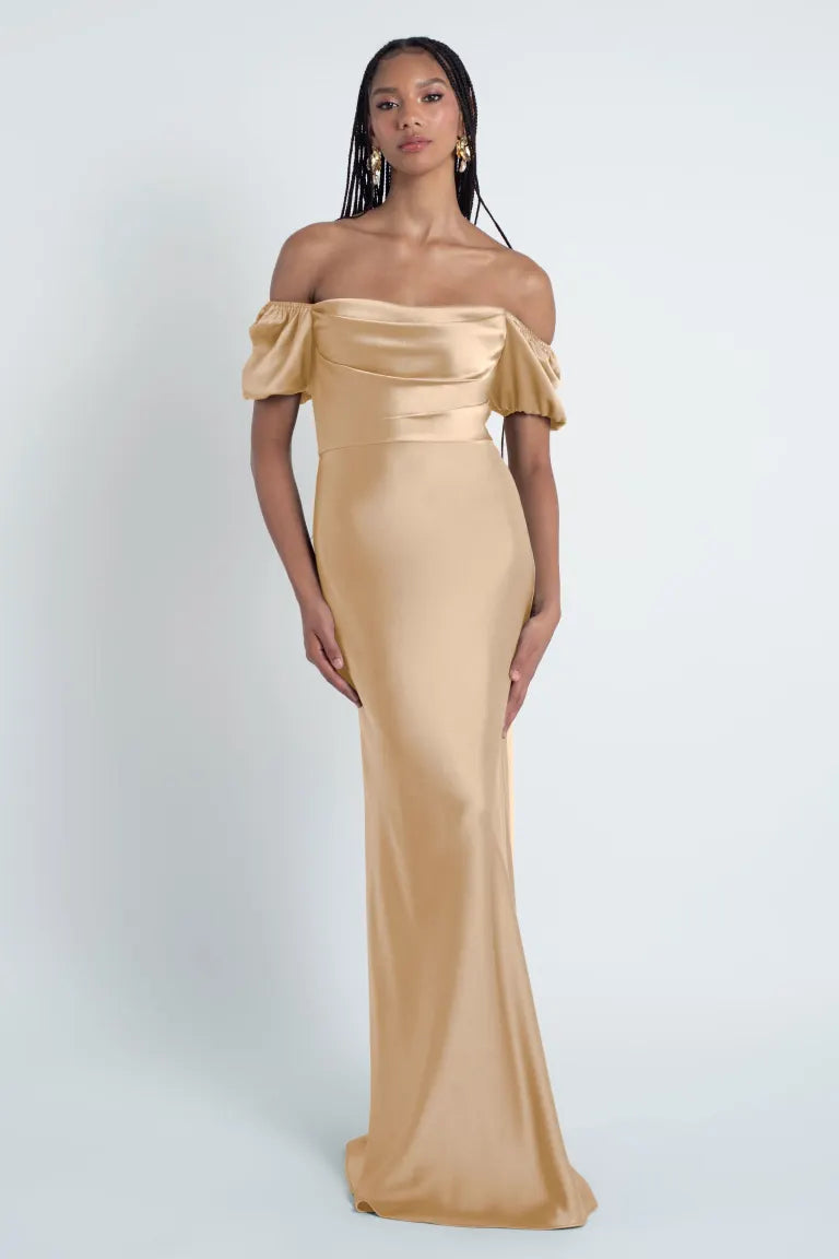 A woman in an elegant off-the-shoulder beige evening gown made of satin fabric, the Eliana - Jenny Yoo Bridesmaid Dress by Bergamot Bridal.