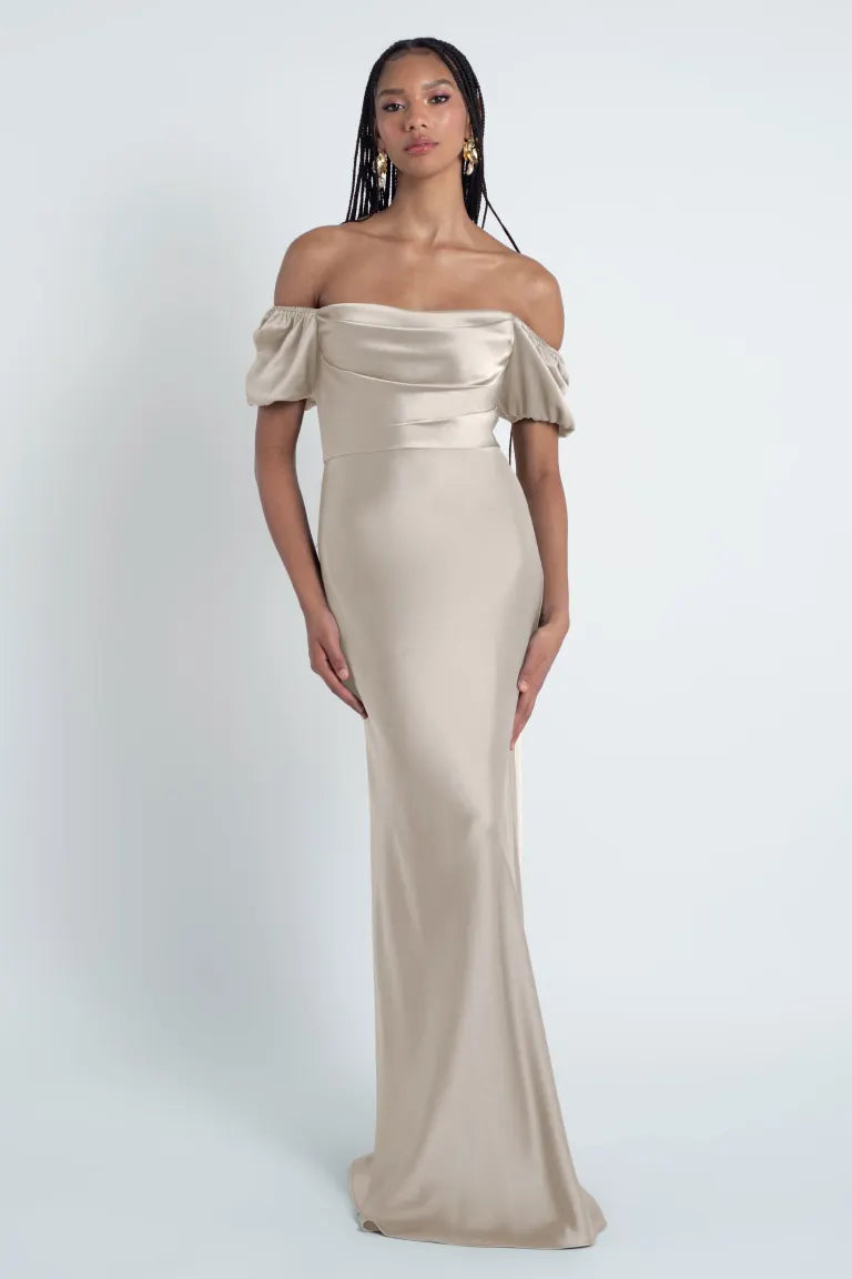 A woman stands posing in an elegant, off-the-shoulder beige gown made of luxe satin fabric by Bergamot Bridal's Eliana - Jenny Yoo Bridesmaid Dress.
