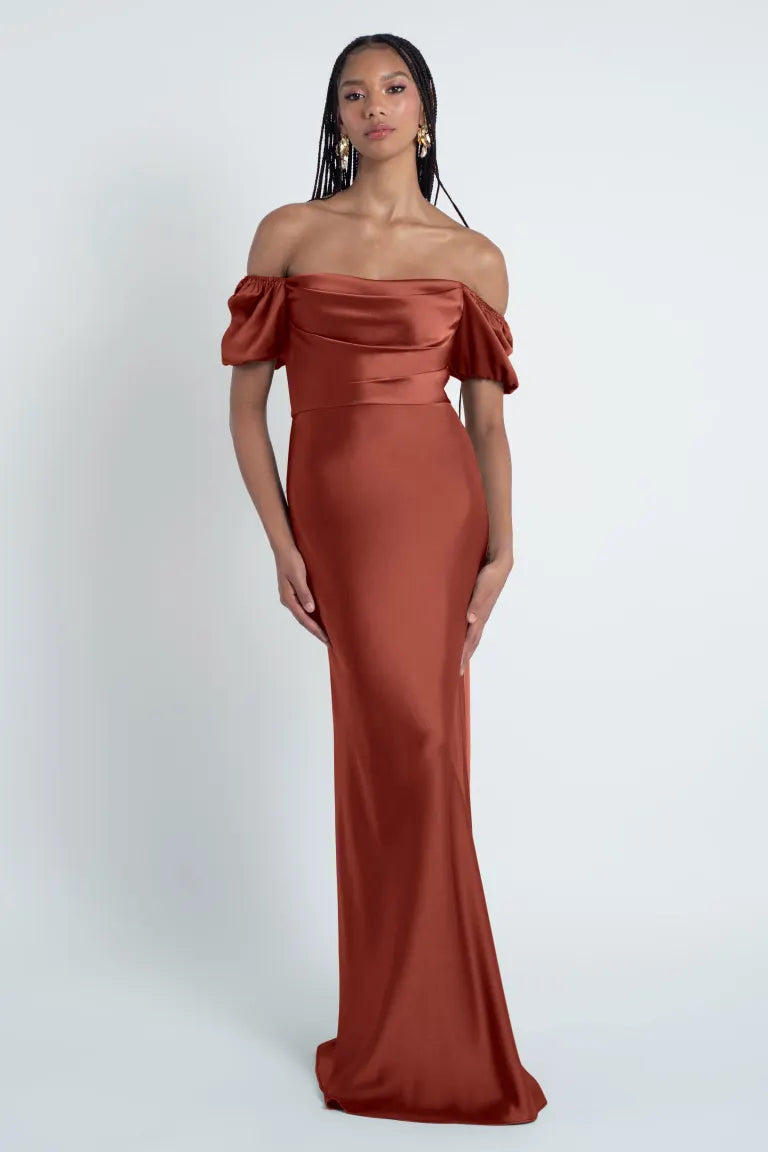 Woman posing in an elegant off-the-shoulder rust-colored Jenny Yoo Bridesmaid Dress made of satin fabric from Bergamot Bridal.