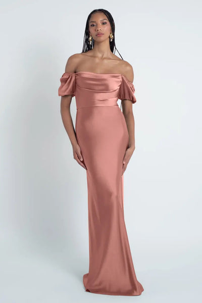 A woman wearing an elegant off-the-shoulder Jenny Yoo Bridesmaid Dress crafted from luxe satin fabric stands against a gray background.