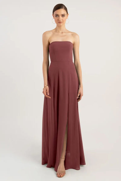 A woman standing against a plain background wearing a Essie - Bridesmaid Dress by Jenny Yoo with a strapless neckline, dusty rose color, and thigh-high slit from Bergamot Bridal.