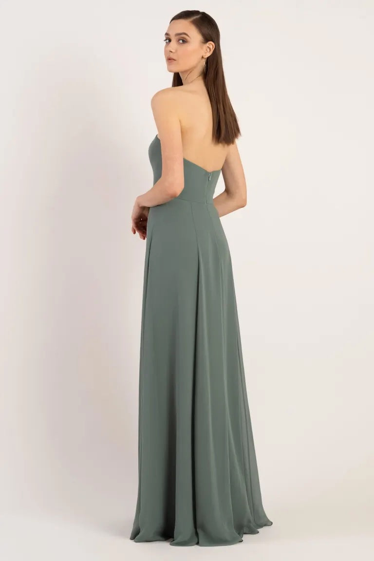 Woman modeling a sleek, backless green evening gown against a neutral backdrop, the Essie - Bridesmaid Dress by Jenny Yoo from Bergamot Bridal.