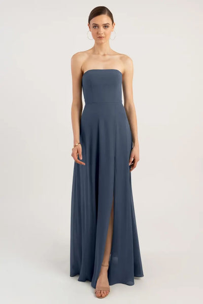 A woman in a strapless Essie - Bridesmaid Dress by Jenny Yoo chiffon bridesmaid dress with a sleek straight neckline and an A-line skirt, featuring a slit, stands against a plain background.