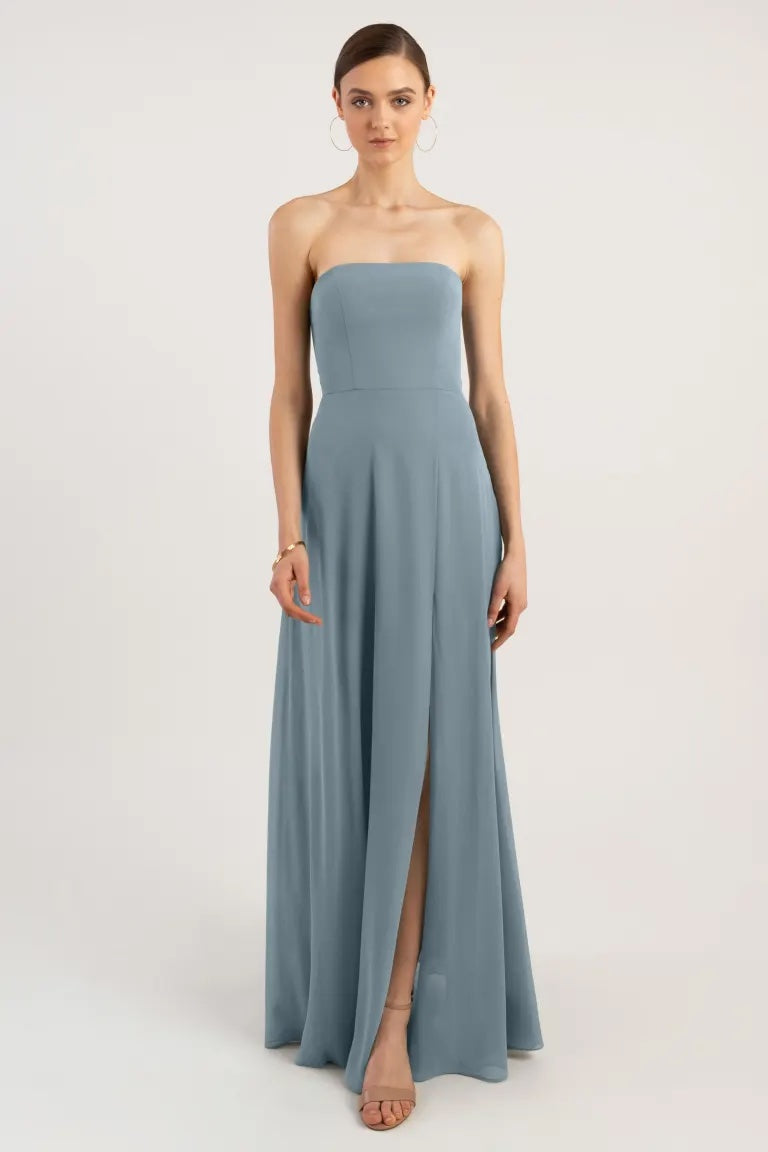 A woman in an Essie - Bridesmaid Dress by Jenny Yoo in a strapless light blue color with a thigh-high slit stands against a plain background from Bergamot Bridal.