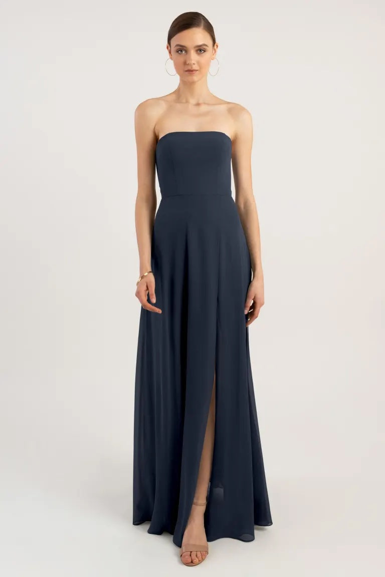 A woman standing against a neutral background wearing a strapless, navy blue bridesmaid dress with a boned bodice and a thigh-high slit, the Essie Bridesmaid Dress by Jenny Yoo from Bergamot Bridal.