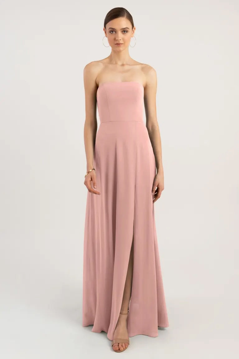 A woman stands wearing an Essie - Bridesmaid Dress by Jenny Yoo in blush pink from Bergamot Bridal with a straight neckline and a thigh-high slit.
