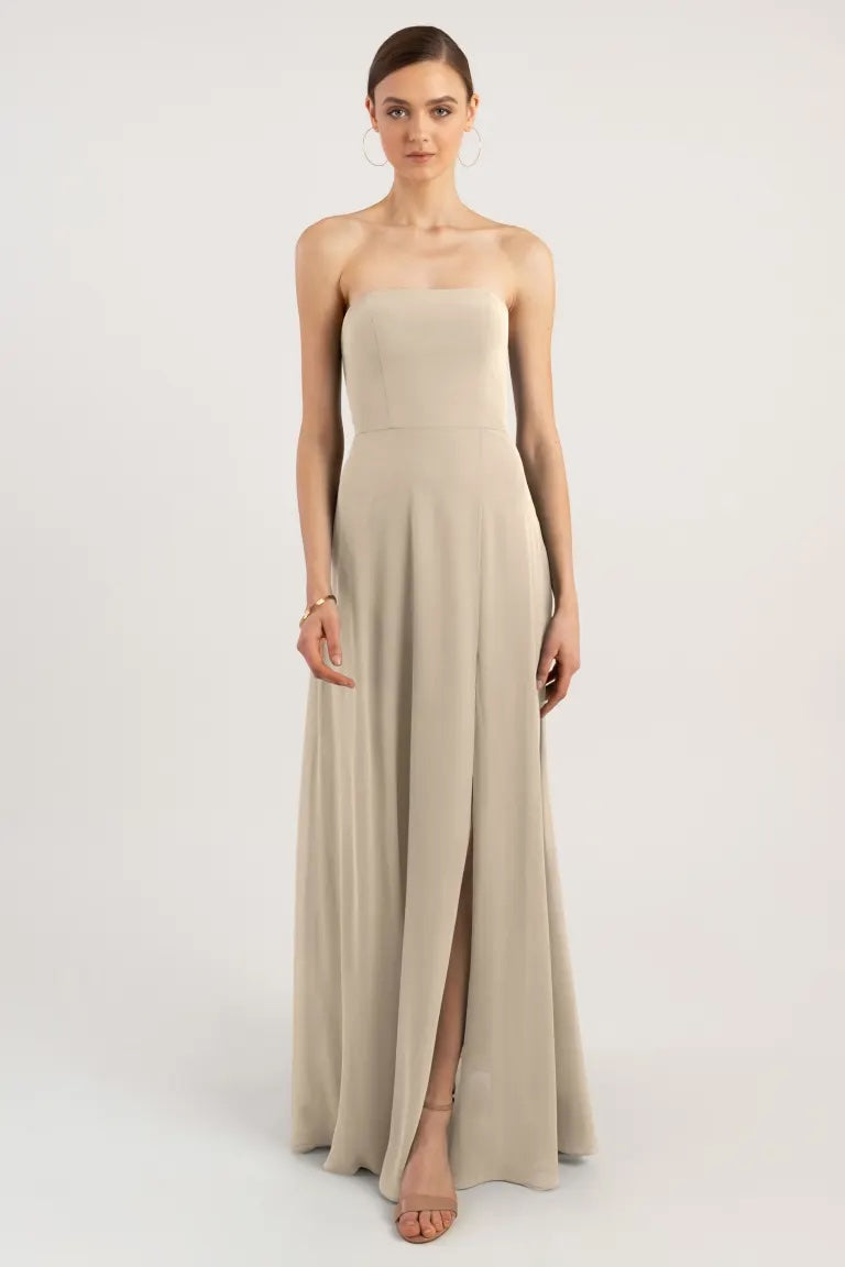 A woman standing against a plain background, modeling a strapless bridesmaid dress with a boned bodice and a slit in the skirt - Essie - Bridesmaid Dress by Jenny Yoo from Bergamot Bridal.