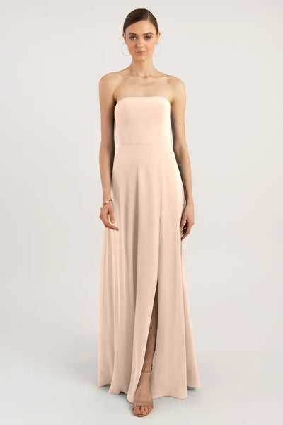 A woman stands wearing a strapless, pale pink evening gown with an A-line skirt and a straight neckline by Bergamot Bridal - Essie - Bridesmaid Dress by Jenny Yoo.
