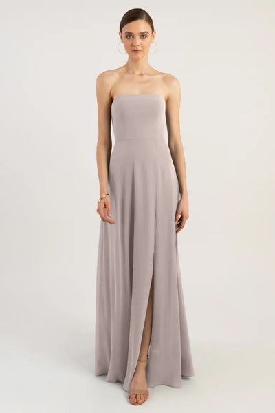 A woman in an elegant strapless neckline taupe Essie bridesmaid dress by Jenny Yoo stands against a neutral background.