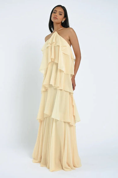 A woman poses in a flowing, cream-colored, sheer chiffon Farley Bridesmaid Dress by Jenny Yoo for Bergamot Bridal against a plain white background.