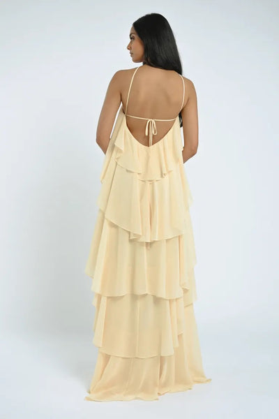 A woman with long dark hair is standing and facing away, wearing the Farley Bridesmaid Dress by Jenny Yoo—a backless, cream-colored chiffon creation with spaghetti straps and a tiered design from Bergamot Bridal—against a plain background, exuding an effortless feel.