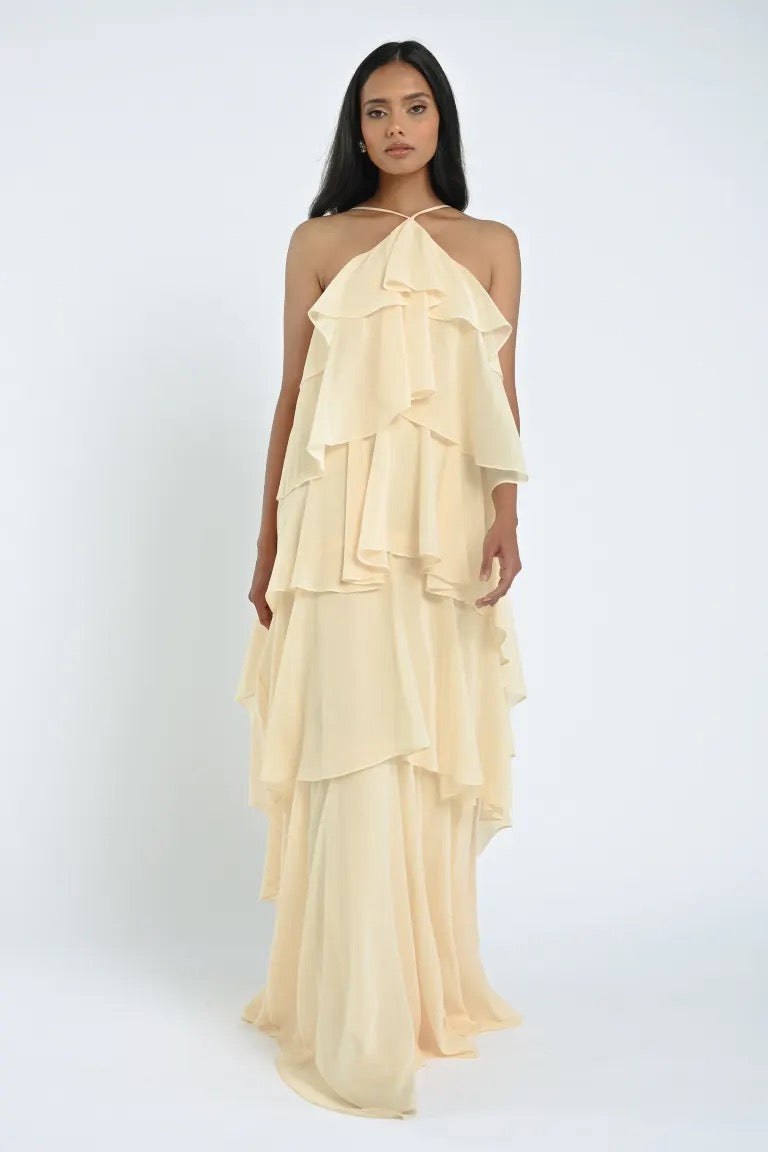 A person stands against a plain background wearing the floor-length, cream-colored, tiered halter Farley Bridesmaid Dress by Jenny Yoo from Bergamot Bridal, exuding an effortless feel.