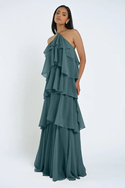 A woman is standing in a floor-length, sleeveless teal chiffon dress. The Farley Bridesmaid Dress by Jenny Yoo from Bergamot Bridal features multiple layered ruffles from the bust to the hem, giving it an effortless feel. She has long, dark hair and is looking straight ahead.