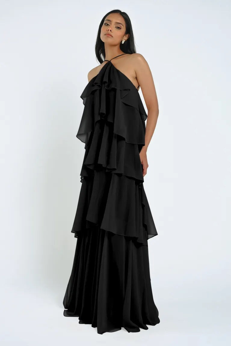 A woman stands wearing the Farley Bridesmaid Dress by Jenny Yoo, a sleeveless, tiered black chiffon gown with a halter neck. She has long dark hair and holds one hand by her side, embodying the effortless elegance of Bergamot Bridal.