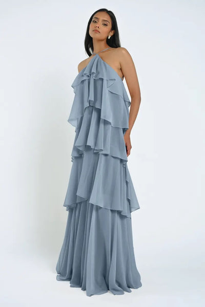 A person wearing a flowing, floor-length Farley Bridesmaid Dress by Jenny Yoo in light blue sheer chiffon with a halter neckline; standing against a plain white background. The dress is from Bergamot Bridal.