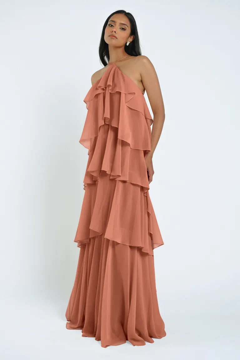 A woman stands wearing the Farley Bridesmaid Dress by Jenny Yoo, a layered, sleeveless, floor-length piece in a muted pink chiffon fabric that exudes an effortless feel against a plain white background. The dress is part of the Bergamot Bridal collection.