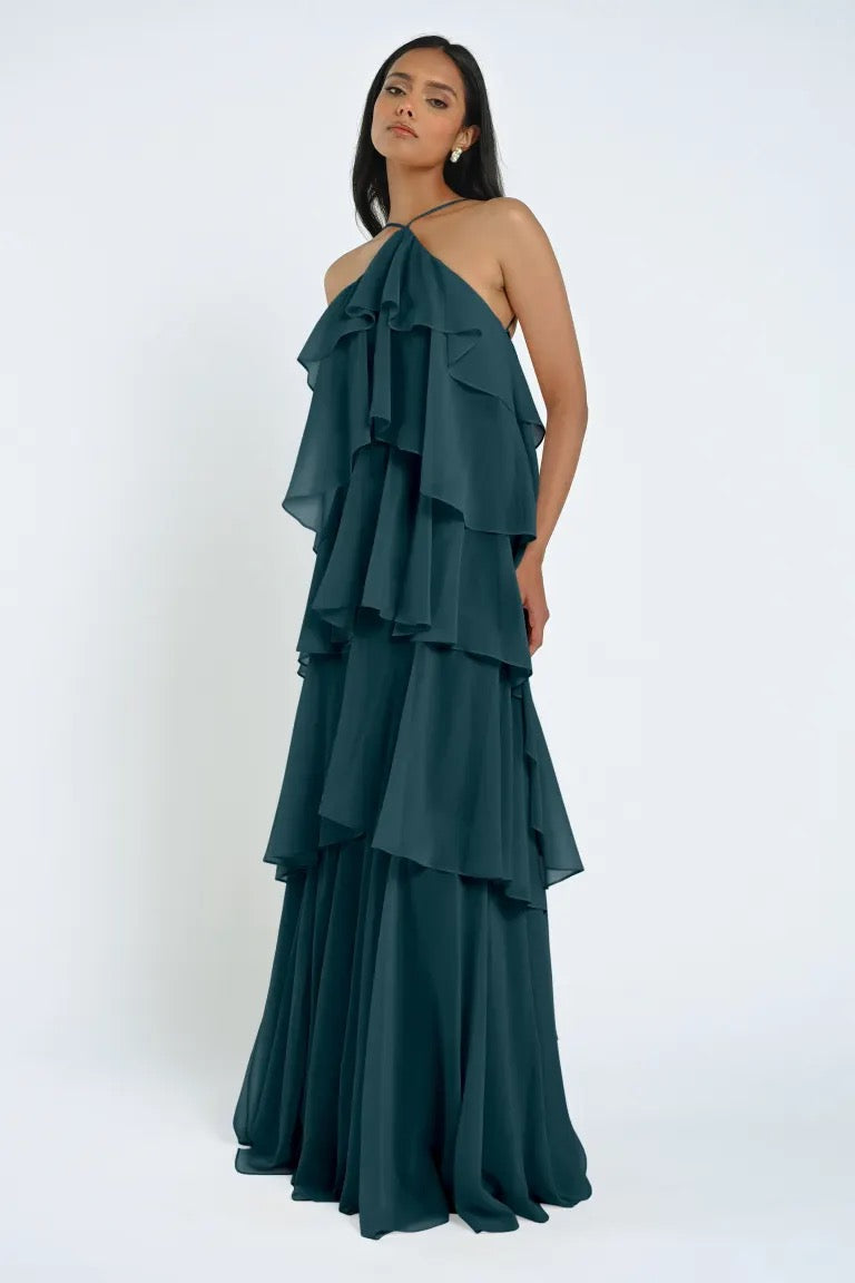 A woman stands in a teal, tiered ruffle halter gown made of chiffon, looking at the camera with a neutral expression against a plain background. The Farley Bridesmaid Dress by Jenny Yoo from Bergamot Bridal exudes an effortless feel, perfectly blending elegance and simplicity.