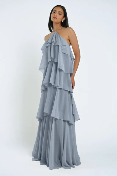 A woman stands wearing a light blue, floor-length Farley Bridesmaid Dress by Jenny Yoo from Bergamot Bridal, featuring cascading chiffon ruffles, set against a plain white background. The airy fabric gives the ensemble an effortless feel.