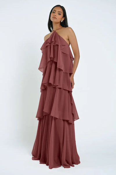 A woman wearing a tiered, ruffled, halter-neck, floor-length mauve Farley Bridesmaid Dress by Jenny Yoo from Bergamot Bridal stands against a plain white background.