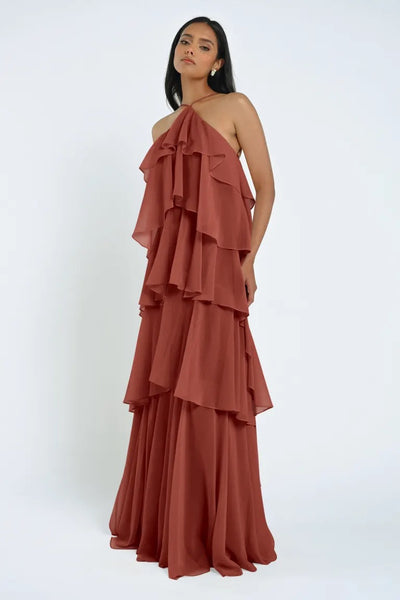 A woman in a tiered, rust-colored Farley Bridesmaid Dress by Jenny Yoo from Bergamot Bridal stands against a plain white background, showcasing the elegant flow of sheer chiffon.