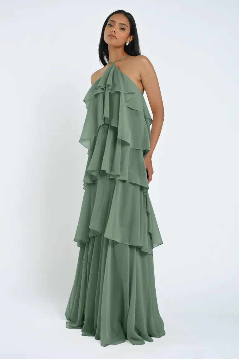 A woman wearing a flowing Farley Bridesmaid Dress by Jenny Yoo in sage green, characterized by its layered halter-neck design, stands against a plain background, exuding an effortless feel with its chiffon fabric. This elegant piece is from Bergamot Bridal.