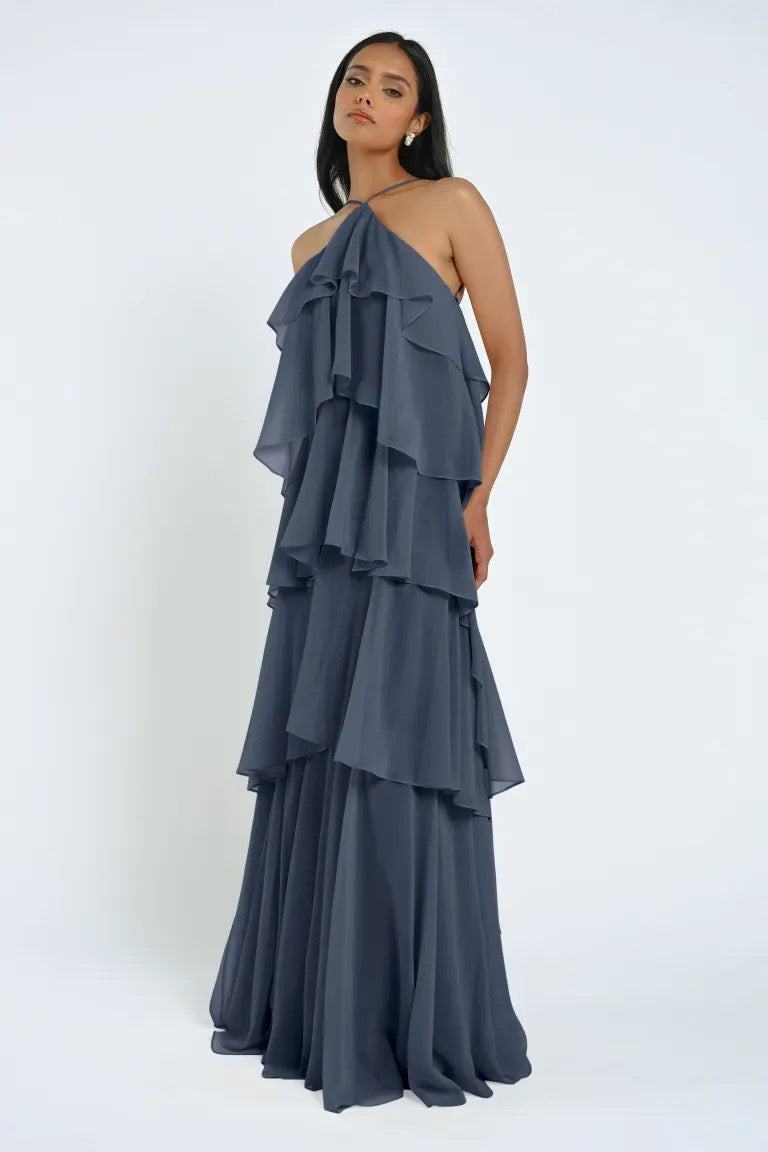 A woman is standing in the Farley Bridesmaid Dress by Jenny Yoo from Bergamot Bridal, a layered, floor-length, halter-neck gray chiffon dress that exudes an effortless feel against a plain background.