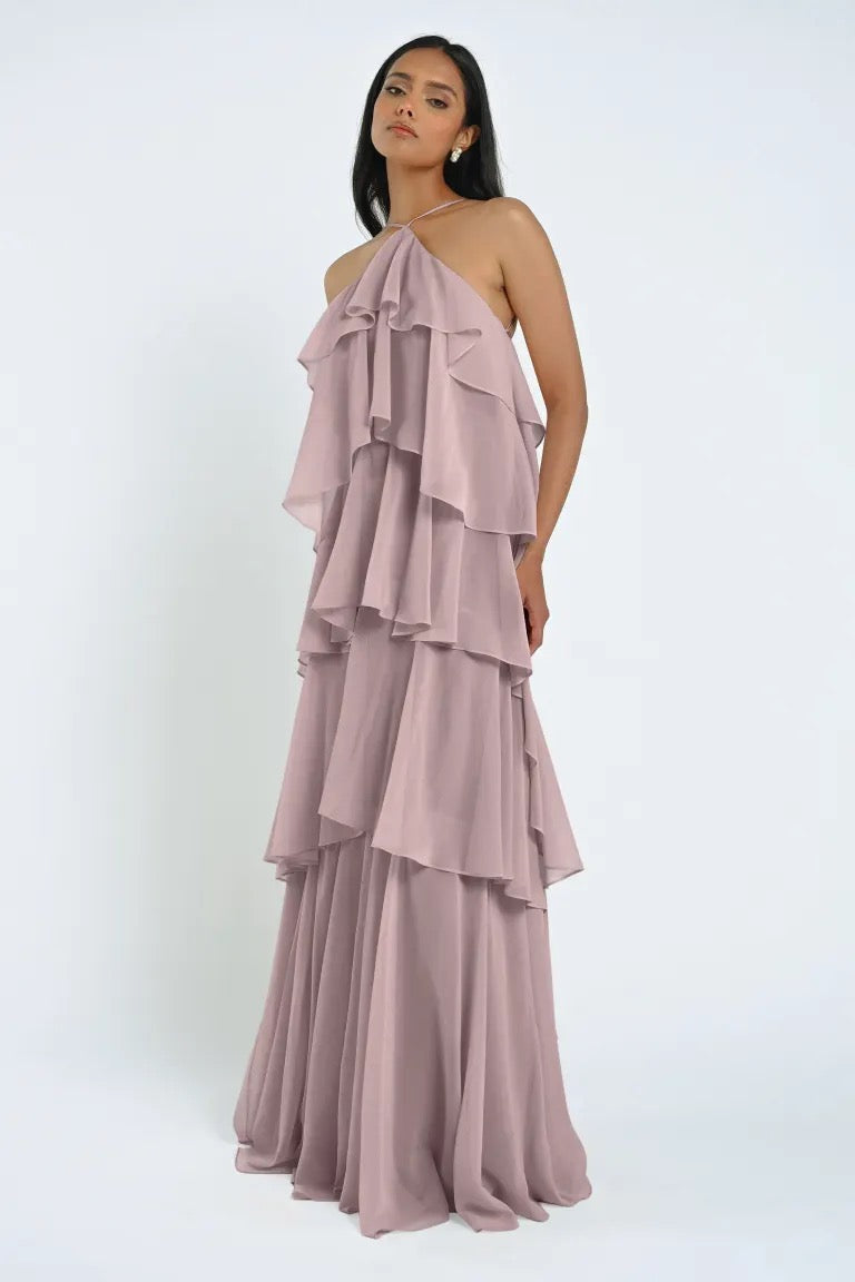 Woman standing against a plain background, wearing the Farley Bridesmaid Dress by Jenny Yoo from Bergamot Bridal—a floor-length, tiered dress with a halter neck in light mauve sheer chiffon.