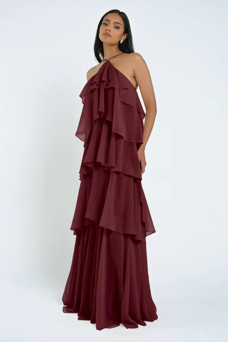 A woman standing in a burgundy, multi-layered chiffon ruffle Farley Bridesmaid Dress by Jenny Yoo with a halter neckline from Bergamot Bridal against a plain white background.