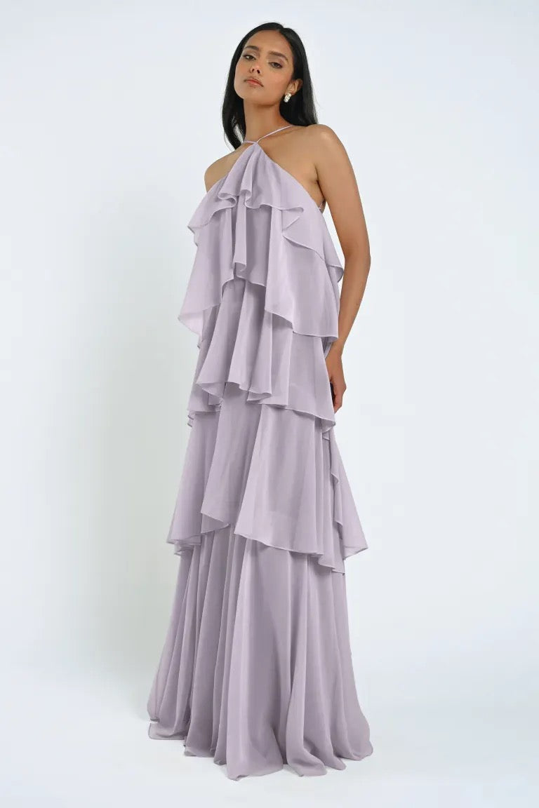 A woman in a flowing, sleeveless lavender-tiered ruffle gown stands against a plain white background, maintaining a neutral expression, effortlessly exuding elegance in the chiffon Farley Bridesmaid Dress by Jenny Yoo from Bergamot Bridal.