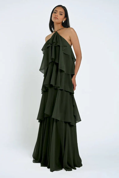 A woman stands wearing the Farley Bridesmaid Dress by Jenny Yoo, a dark green, tiered halter gown with ruffled layers of chiffon from Bergamot Bridal. With her arms relaxed at her sides and looking directly at the camera, she exudes an effortless feel against the plain light grey background.