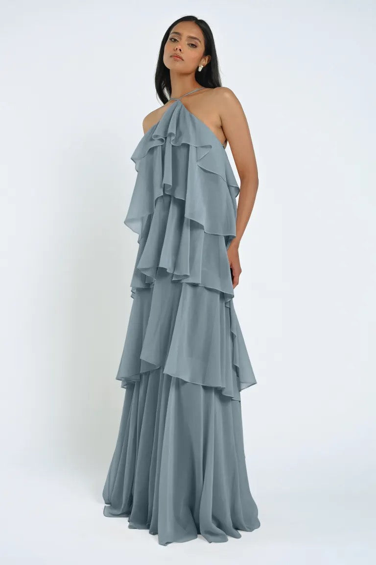 A woman in a tiered, light blue Farley Bridesmaid Dress by Jenny Yoo from Bergamot Bridal stands against a plain white background. She has long dark hair and a neutral expression.