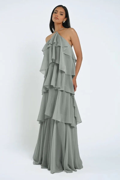 A woman wearing an elegant, tiered grey Farley Bridesmaid Dress by Jenny Yoo from Bergamot Bridal stands against a plain white background.