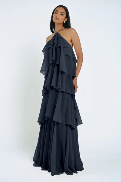 A woman wearing a Bergamot Bridal Farley Bridesmaid Dress by Jenny Yoo in navy blue, featuring a tiered design and halter-neck, stands against a plain white background, looking directly at the camera and exuding an effortless feel.