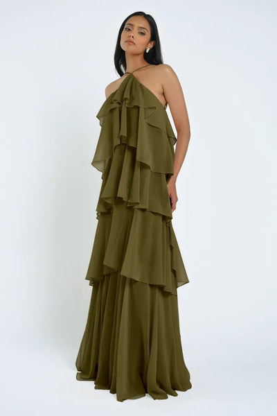 A woman is standing against a plain background wearing an olive green Farley Bridesmaid Dress by Jenny Yoo from Bergamot Bridal. The dress features multiple ruffled layers, giving it an effortless feel. The halter-neck style and chiffon fabric add elegance as she rests one hand on her hip.