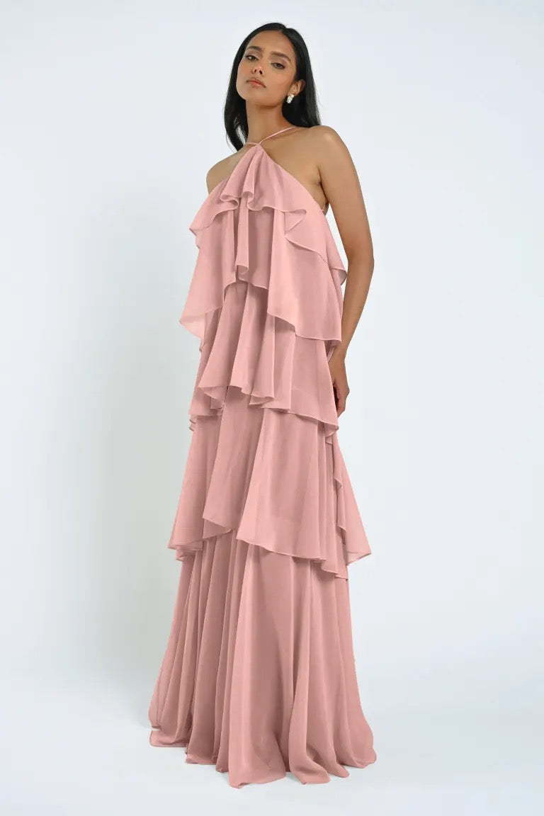 A woman in a long, pink Farley Bridesmaid Dress by Jenny Yoo from Bergamot Bridal with tiered ruffle layers poses against a plain white background.