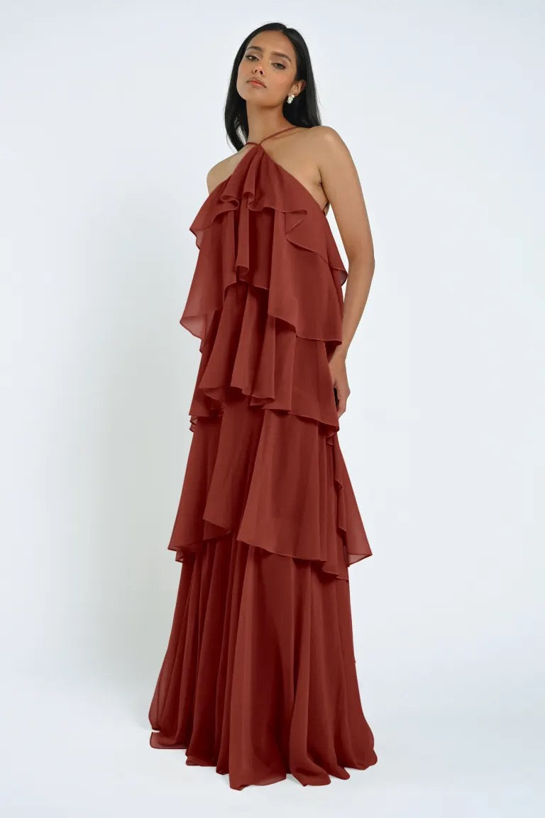 Woman standing against a plain background wearing a rust-colored, tiered, halter-neck Farley Bridesmaid Dress by Jenny Yoo from Bergamot Bridal, exuding an effortless feel.