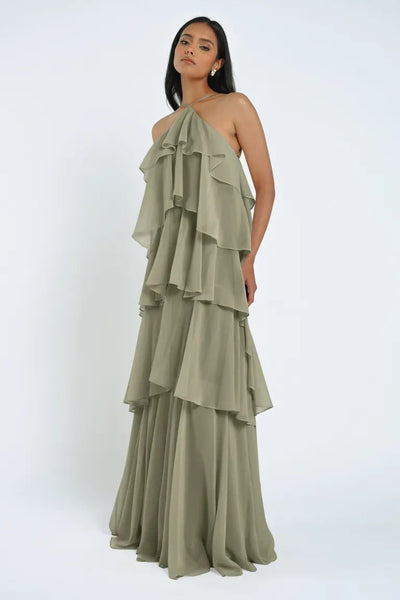 A woman is standing in a tiered, sleeveless sage green chiffon Farley Bridesmaid Dress by Jenny Yoo from Bergamot Bridal that drapes elegantly to the floor, exuding an effortless feel. She has long, dark hair and a neutral expression against a plain white background.