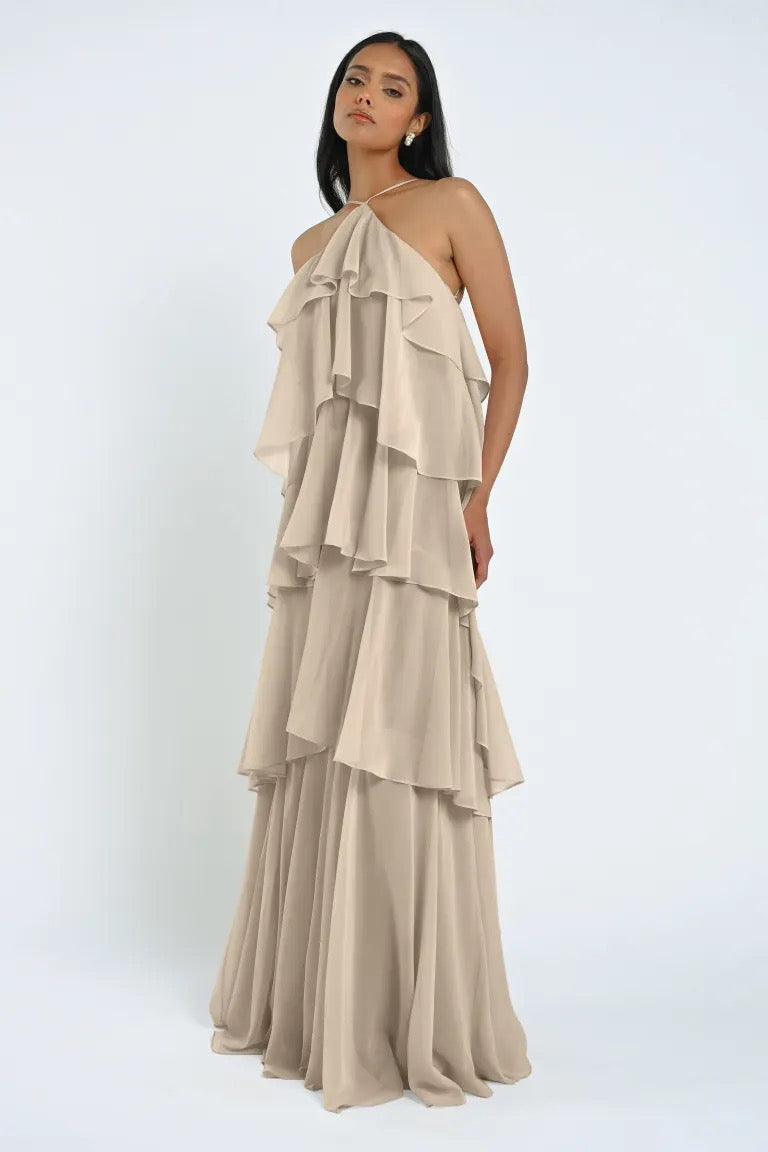 A person stands wearing the tiered, beige halter-neck Farley Bridesmaid Dress by Jenny Yoo from Bergamot Bridal, made of sheer chiffon against a plain white background.